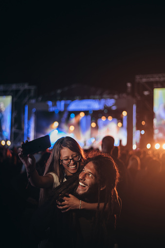 Teenage couple at summer music festival taking selfie with a smartphone while dancing in front of stage. New music entertainment trends. People in love.