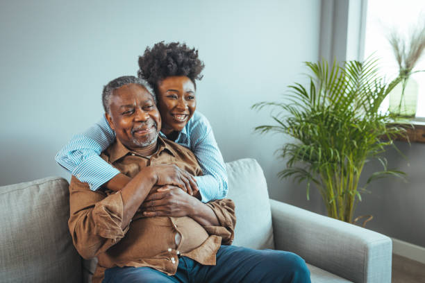 Senior man and his middle aged daughter smiling at each other embracing, close up. Senior man and his middle aged daughter smiling at each other embracing, close up. Portrait of a daughter holding her elderly father, sitting on a bed by a window in her father's room. father and daughter stock pictures, royalty-free photos & images