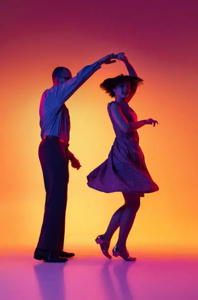 Photo of Portrait of excited man and woman, couple of dancers in vintage retro style outfits dancing lindy hop dance isolated on gradient yellow and purple background.