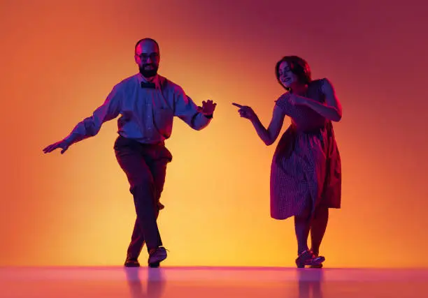 Photo of Portrait of excited man and woman, couple of dancers in vintage retro style outfits dancing lindy hop dance isolated on gradient yellow and purple background.