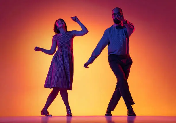 Photo of Portrait of excited man and woman, couple of dancers in vintage retro style outfits dancing lindy hop dance isolated on gradient yellow and purple background.
