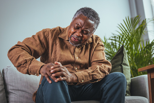 Photo of mature, elderly man sitting on a sofa in the living room at home and touching his knee by the pain during the day. Mature man massaging his painful knee.