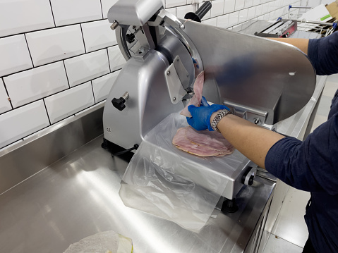 Hands of a shop assistant wearing disposable plastic gloves using ham slicer machine.