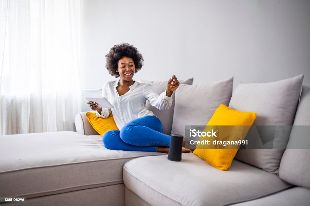 Black woman connecting phone to her virtual assistant smart speaker. Smart wireless column with a young woman controlling home devices with a voice commands on the background at home. Woman controlling home devices with a voice commands Virtual Assistant Stock Photo