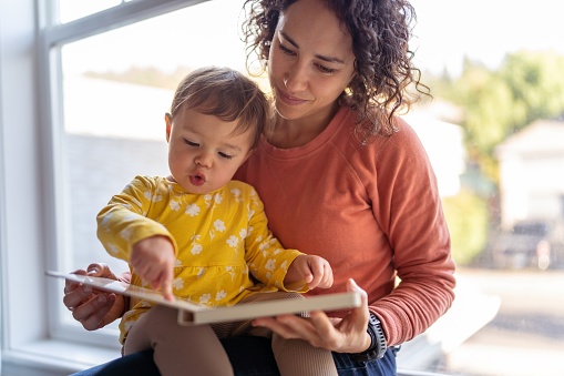A loving mother of Pacific Islander descent sits on the windowsill and reads a storybook to her preschool age daughter. The adorable Eurasian child is sitting on her mother's lap and is pointing with curiosity at the book's pages.