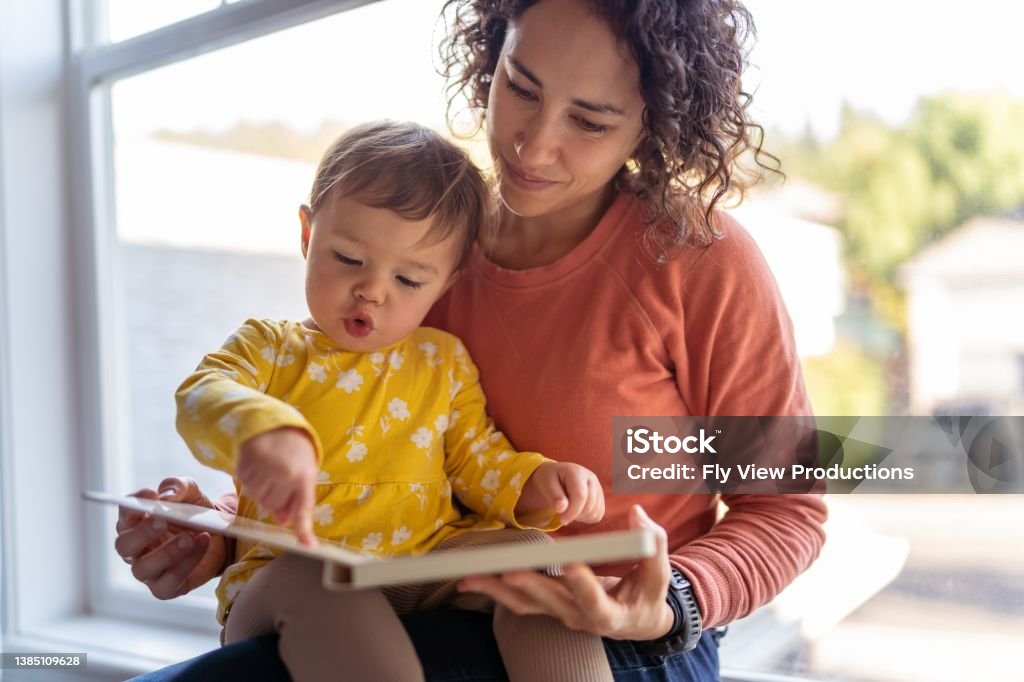 Affectionate mother reading book with adorable toddler daughter A loving mother of Pacific Islander descent sits on the windowsill and reads a storybook to her preschool age daughter. The adorable Eurasian child is sitting on her mother's lap and is pointing with curiosity at the book's pages. Toddler Stock Photo