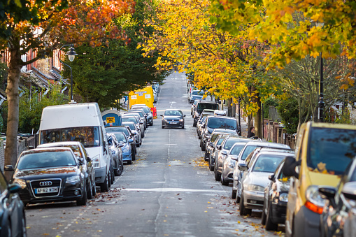 London, UK - 25 October, 2021 - A car driving through a treelined street with cars parked on both sides during autumn season in Crouch End area