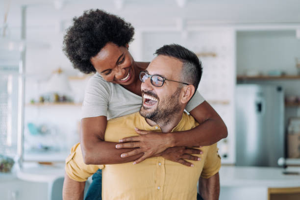 Young happy couple piggybacking at home. Shot of young couple spending leisure time together at home. Smiling girl piggybacking on her boyfriend. Playful man having fun while piggybacking his girlfriend. Happy man giving piggyback ride to woman in the living room. 30s 40s activity adult stock pictures, royalty-free photos & images