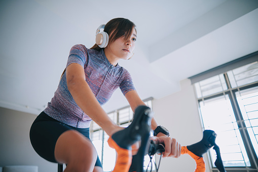 Asian Chinese woman cycling with stationary bicycle trainer and listening to music with headphone at home