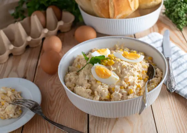 Traditional savory tuna salad cooked with boiled eggs and marinated with mayonnaise, onions and corn. Served in a white bowl on wooden kitchen table. Ready to eat