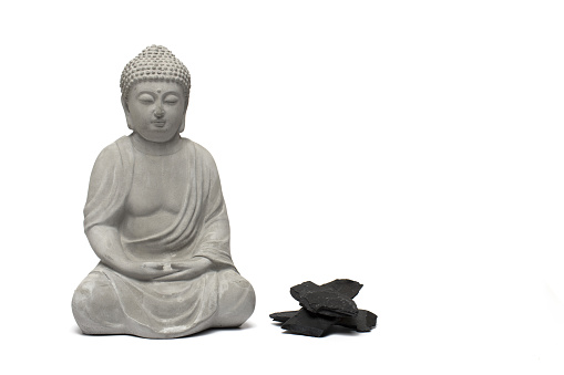 Close up of cast sitting buddha figure next to dark stacked stones on white background
