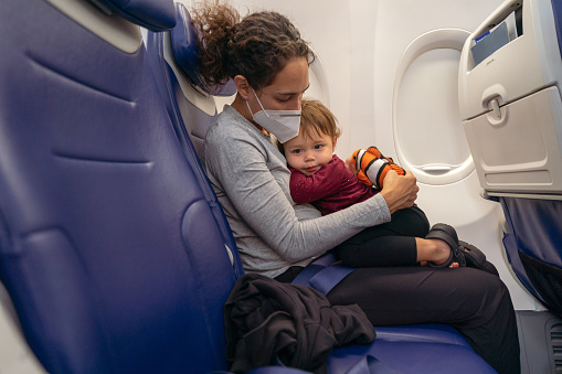 A young mom of Pacific Islander descent wearing a protective face mask affectionately holds her toddler daughter on her lap while traveling on an airplane.