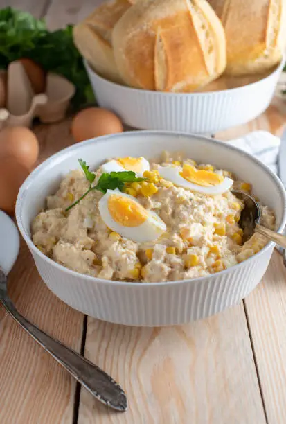Traditional homemade savory tuna salad with mayonnaise, boiled eggs, onions and corn. Served in a white bowl with spoon and buns on wooden table. Ready to eat. Vertical image