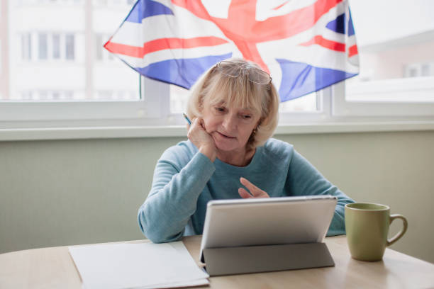 Senior woman learns English via internet with laptop, language classes for Ukrainian refugee Senior woman sits at desk before laptop and learns English via internet, language classes for Ukrainian refugee for better adaptation abroad, British flag on the window, social distance education Teaching English Abroad stock pictures, royalty-free photos & images