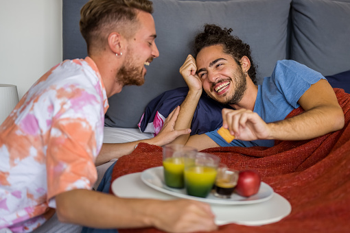 Young gay couple having breakfast at home in bed just woke up, focus on the toothy smile of the man lying in bed, organic breakfast with biological juice and fruits