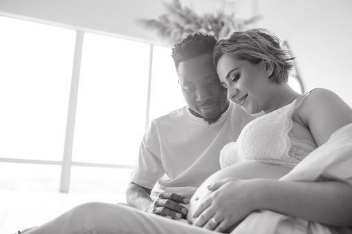 Young happy african man and his caucasian pregnant woman and sit and touch her belly by their hands. Interracial marriage concept. Black and white image.