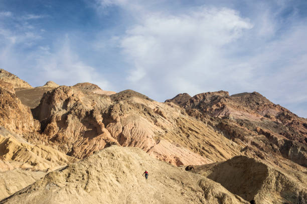 istock Man Climbs Rugged Desert Mountains with Blue Sky and Clouds Landscape 1385156198