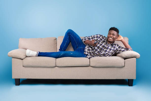 Weekend leisure activities. Millennial black man with remote control switching channels on TV while lying on couch