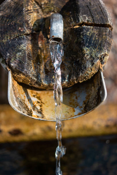 istock Old rustic drinking water fountain in the forest. Wooden construction, rusty metal pipe and faucet, streaming water. Close up shot, shallow depth of field, no people 1385175061