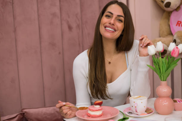 Pretty smiling woman eating delicious sweet pink macaroons, stock photo Woman in white shirt, eating delicious sweet pink macaroons with a strawberry on the top, next to cup a coffee/ cappuccino, in a decorated room cleavage stock pictures, royalty-free photos & images