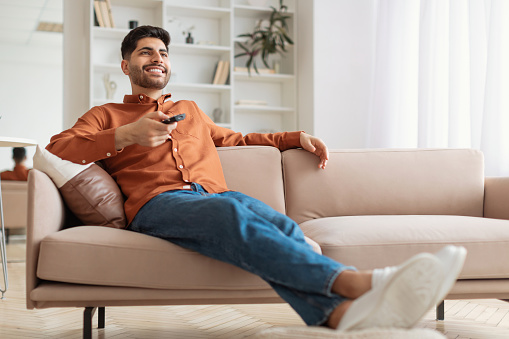 Portrait of Arab guy watching TV show or film, holding remote control, switching channels. Young smiling man spending weekend free time sitting on comfortable sofa at home in living room, copy space