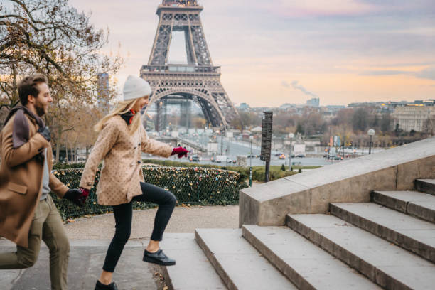 Couple, man with short brown hair and beard, wearing a beige coat, woman with long blonde hair wearing a white hat and beige jacket with brown dots, holding hands while smiling and running up stairs to Parvis des Droits de l'Homme observation platform in front of Eiffel Tower, Paris, street with traffic in the background, twilight, side view, horizontal