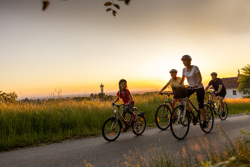 Family, Parents biking with their son and little daughter on a country lane, mother and daughter in the front, daughter is looking at her mother, all of them are smiling, wearing short summer clothes and helmets, in the evening with wheat field and sunset in background, front view, horizontal