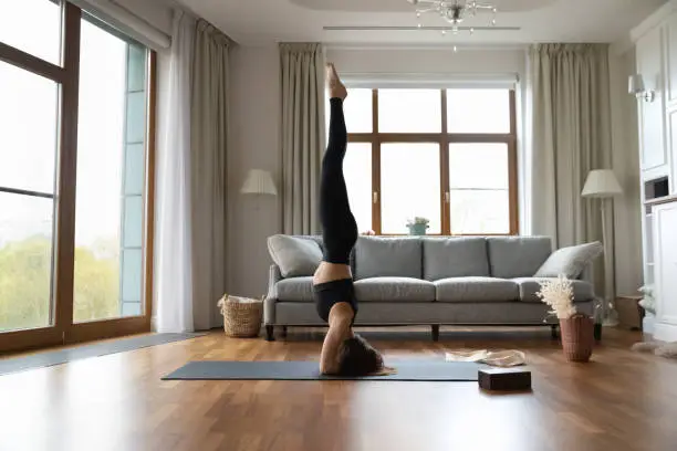 Slim sporty woman wear black sportswear working out alone in living room, performs supported headstand or Salamba Sirsasana exercise, side full length view. Yoga asanas practice, sport at home concept