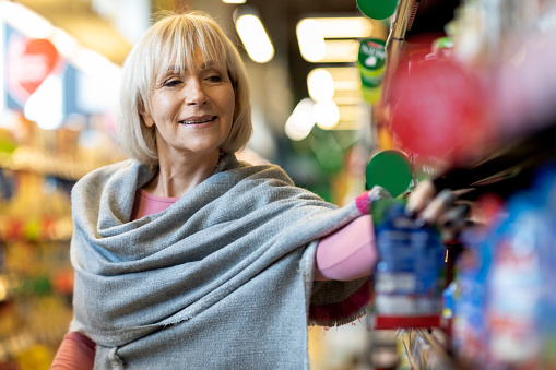 Closeup of attractive elderly lady with shawl on her shoulders making shopping at supermarket, standing in store aisle, picking up quality goods from shelves, panorama with copy space
