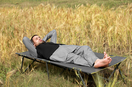 Rural farmer of Indian ethnicity lying down on camp bed near green field during springtime.