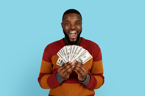 Big Profit. Excited African American Man Holding Lots Of Dolar Cash In Hands And Exclaiming With Joy, Happy Cheerful Black Male Standing With Money Fan Over Blue Studio Background, Copy Space