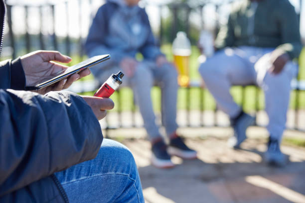 close up of teenagers with mobile phone vaping and drinking alcohol in park - vape stockfoto's en -beelden