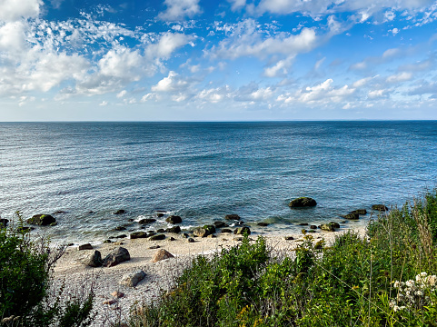 Calm waters of Long Island Sound, with large rocks strewn on sandy beach, blue sky with fluffy clouds and a bit of grassy dune in foreground.