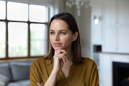 Head shot portrait pensive beautiful woman touch chin staring into distance looks thoughtful. Pretty housewife deep in thoughts standing alone in modern living room consider idea of house remodelling