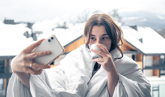 A young woman with a cup of coffee on the balcony in the hotel takes a selfie in the morning, wrapped in a blanket, in the winter in the mountains.