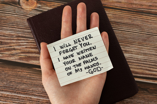 Human hand holding a handwritten Bible quote about God Jesus Christ's love and faithfulness on a wooden background. Top view. A closeup.