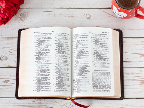 Open Holy Bible Book with a cup of coffee, and red flowers on a wooden table. Top view. Biblical concept of morning prayer, devotion, faith, and study from Scriptures inspired by God Jesus Christ.