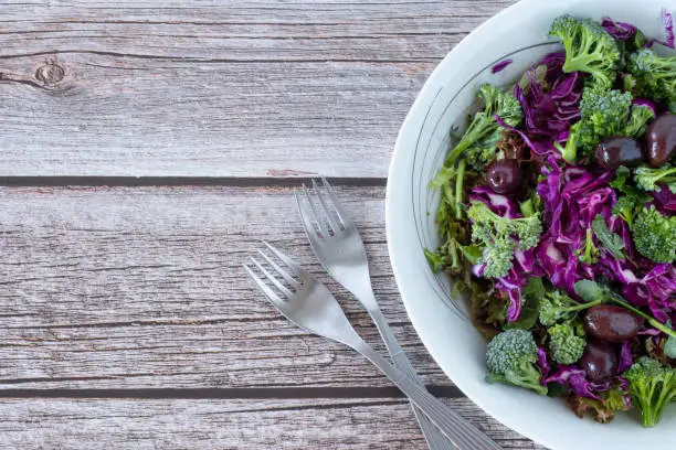 Fresh colorful vegetable salad bowl with red cabbage, broccoli, olives, and lettuce with forks on wooden table with copy space. The concept of healthy plant-based eating.