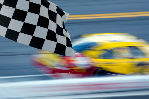 race car blurred on track against rail, checkered flag in foreground, no people, sunny day, car in motion
