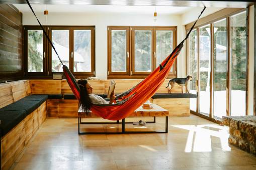 Woman in her late 30s reading a book in the mountain cabin, relaxing in the hammock next to the fireplace.