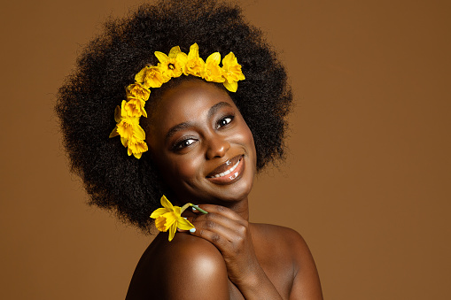 Black Woman Beauty with Yellow Narcissus Flower. African Model with Curly Coil Hairstyle and Floral Wreath in Hair over Beige Studio Background. Face Skin and Body Care