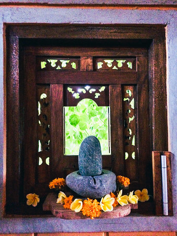 Vertical closeup photo of a small devotional altar in a Balinese family shrine with a carved granite Lingam and Yoni bowl surrounded by fresh yellow Frangipani and orange Marigold flowers on a recessed windowsill with carved wooden shutters. Ubud, Bali 2017