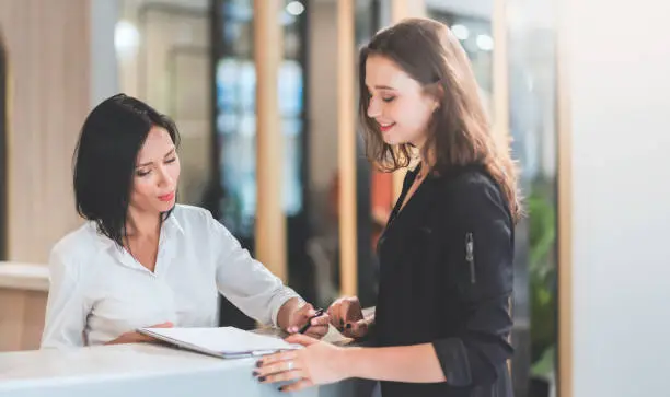 Beautiful receptionist woman talking and showing rates of their services to female client at reception of hotel or office, reservation and check in