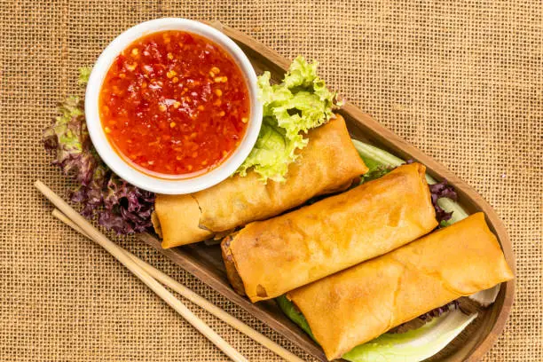 Top view of delicious homemade deep fried spring rolls in wooden tray with red chili sweet sauce in white ceramic cup and green vegetables.
