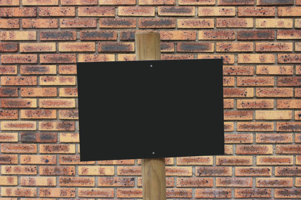 istock Blank black colored information board attached to a wooden pole  Brick wall background 1385337792