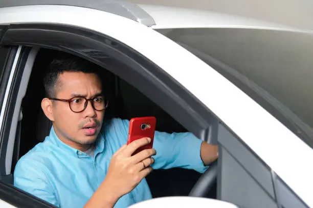 Photo of Adult Asian man looking to his mobile phone with serious expression while driving a car