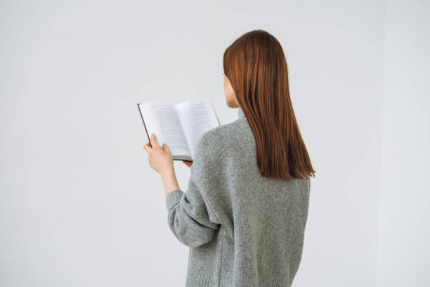 красивая молодая женщина с темными длинными волосами в сером вязаном свитере читает книгу на белом фоне - book reading white women стоковые фото и изображения
