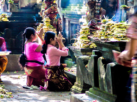 Horizontal photo of two Balinese women, dressed in brightly colored temple clothing, kneeling on the ground praying in front of a religious statue in the Central Market temple, Ubud, Bali. Jalan Raya, Ubud, Bali, Indonesia.