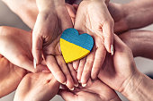 Wir stehen mit dem Symbol der Ukraine, da sich zahlreiche Hände in Einheit mit einem holzfarbenen Herzen in ukrainischen Flaggenfarben halten.