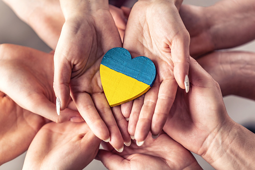 We stand with Ukraine symbol as numerous hands hold one another in unity with a wooden heart painted in Ukrainian flag colors on top.